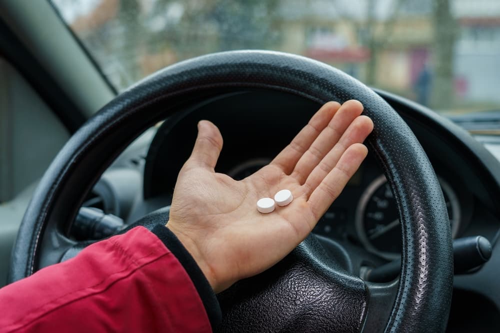 A person holding two pills in their hand while sitting behind a car's steering wheel.